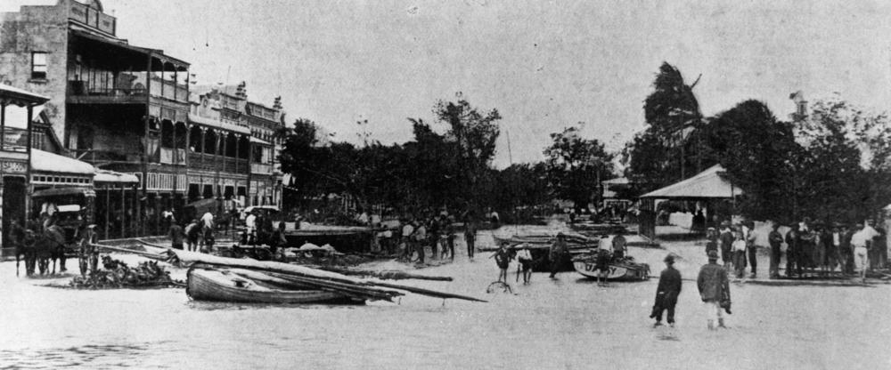 Tidal wave waters in Abbott Street, Cairns, after cyclone, showing boats sheltering in temporary havens. People congregate in the street and wade through the waters. A horsedrawn carriage passes in front of the shops on the left. Some tree-damage is seen on the right`.  'John Oxley Library, State Library of Queensland Image: 158092'.