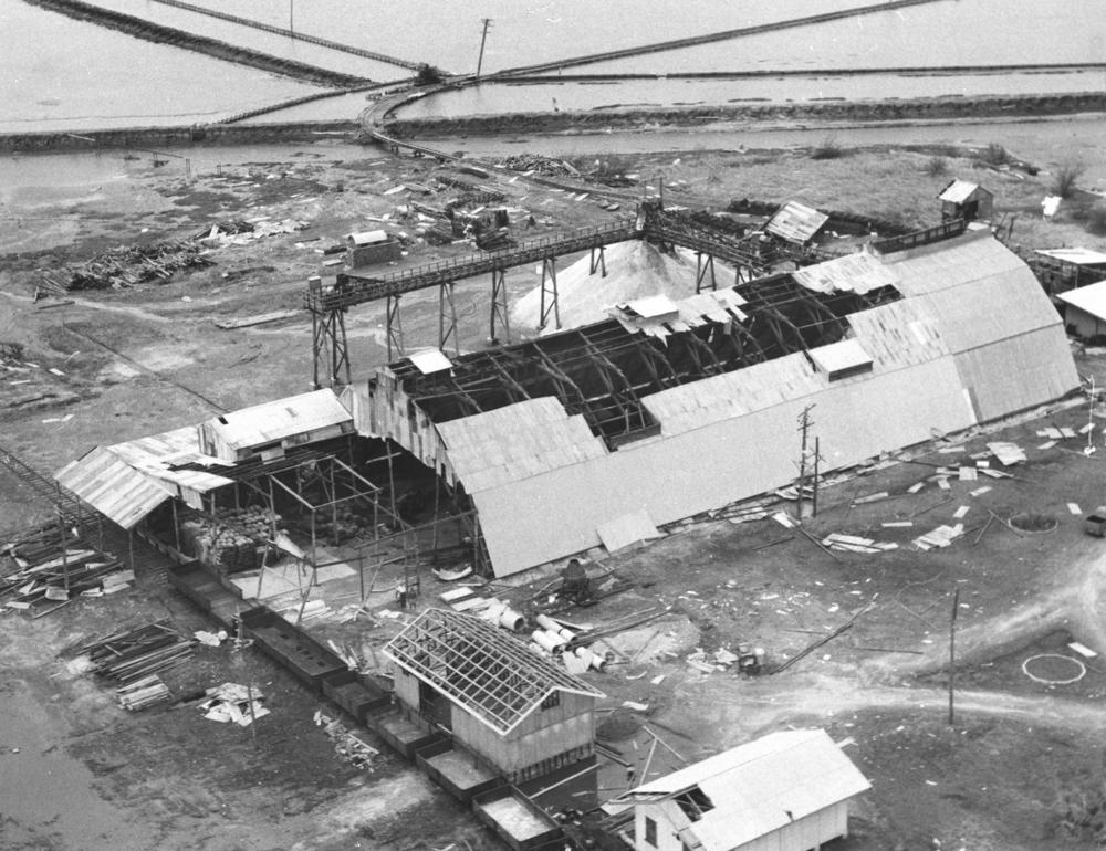 Cyclone Connie caused severe damage to many buildings in Bowen on 16 February 1959. The saltworks building has lost most of the roof and debris is scattered all around the exterior. A small building next to the larger building has lost the whole roof. 'John Oxley Library, State Library of Queensland Image: 201247'.