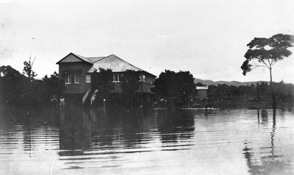 Two people stand in front of a highset Queenslander house during a flood at Tully. The water is knee-high.  'John Oxley Library, State Library of Queensland image: 41491'