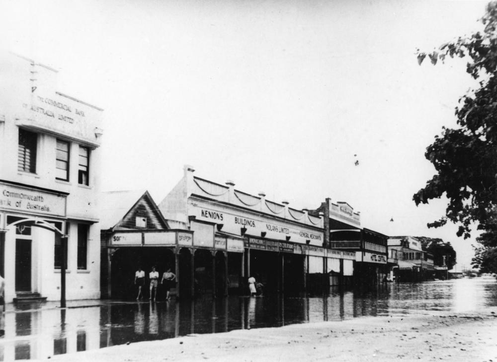 Floodwaters sweep down in front of the Kenions Building in Ingham. Adjoining businesses including the Hotel Central and Nolans Limited have water covering footpaths in front of the shops.The Commonweatlth Bank of Australia has less water damage. 'John Oxley Library, State Library of Queensland image: 24661'