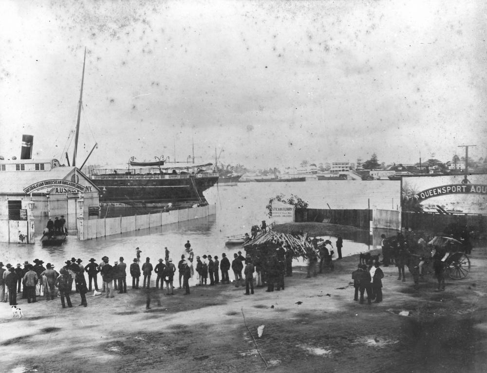 Men are lined up along the waters edge opposite the flooded entrance to the Australasian United Steam Navigation Company wharf where a boat can be seen ferrying people inside. A large steam ship is docked along the wharf. Opposite the wharf is the steam ferry dock together with a large advertisment for the Queensport Aquarium. Scene near the intersection of Creek Street and Eagle Streets, with the AUSN wharves at left, during the floods of 22-25 January 1887. 'John Oxley Library, State Library of Queensland Image: 66442'.