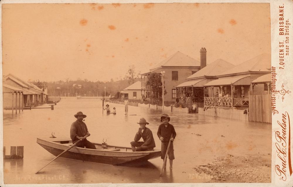 View of flooded street scene at Breakfast Creek, Brisbane, 1890. A man and a boy sit in a rowboat in the foreground and a boy stands next to them. Other people stand in the water or on the verandahs of the houses to the right. The fronts of some houses are visible on the left. The houses on the bush-clad hill across the river can be seen in the background . 'John Oxley Library, State Library of Queensland   Image:60383'.